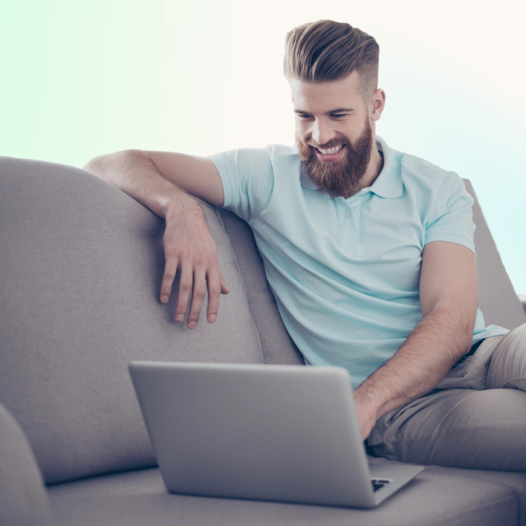 Smiling middle-aged bearded man with pale skin and tidy light brown hair sits on couch looking at laptop in relaxed position.